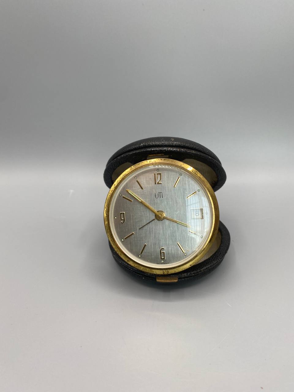 Pendulette de voyage UTI France, cadran guillouché argenté avec calendrier, étui cuir noir ouvert