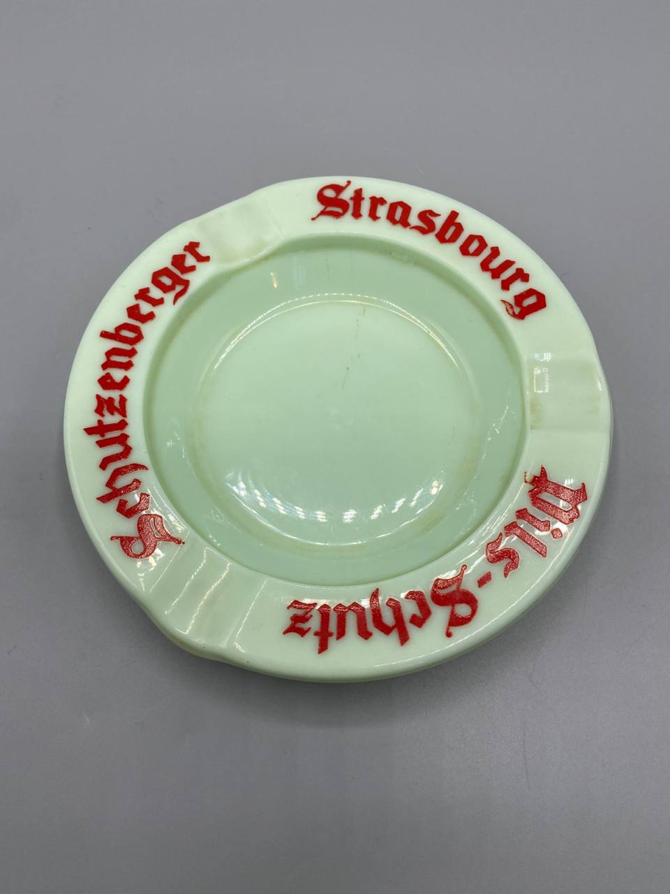 Cendrier publicitaire Schutzenberger Strasbourg, vue de dessus, verre opalin vert pâle, inscription gothique rouge Schutzenberger Strasbourg et Pils-Schutz