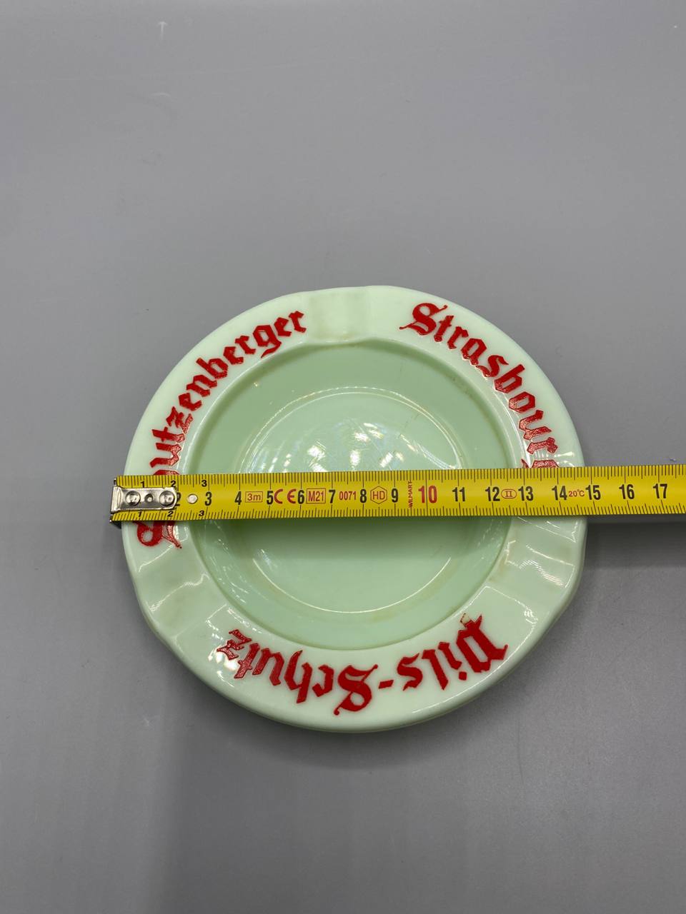Cendrier Schutzenberger avec mètre ruban, diamètre environ 15 cm, vue de dessus, inscription rouge visible