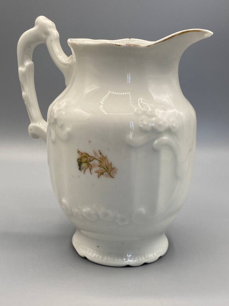 Pot à lait porcelaine, face arrière, panse en relief avec motifs floraux discrets, anse en volute sculptée, pied festonné