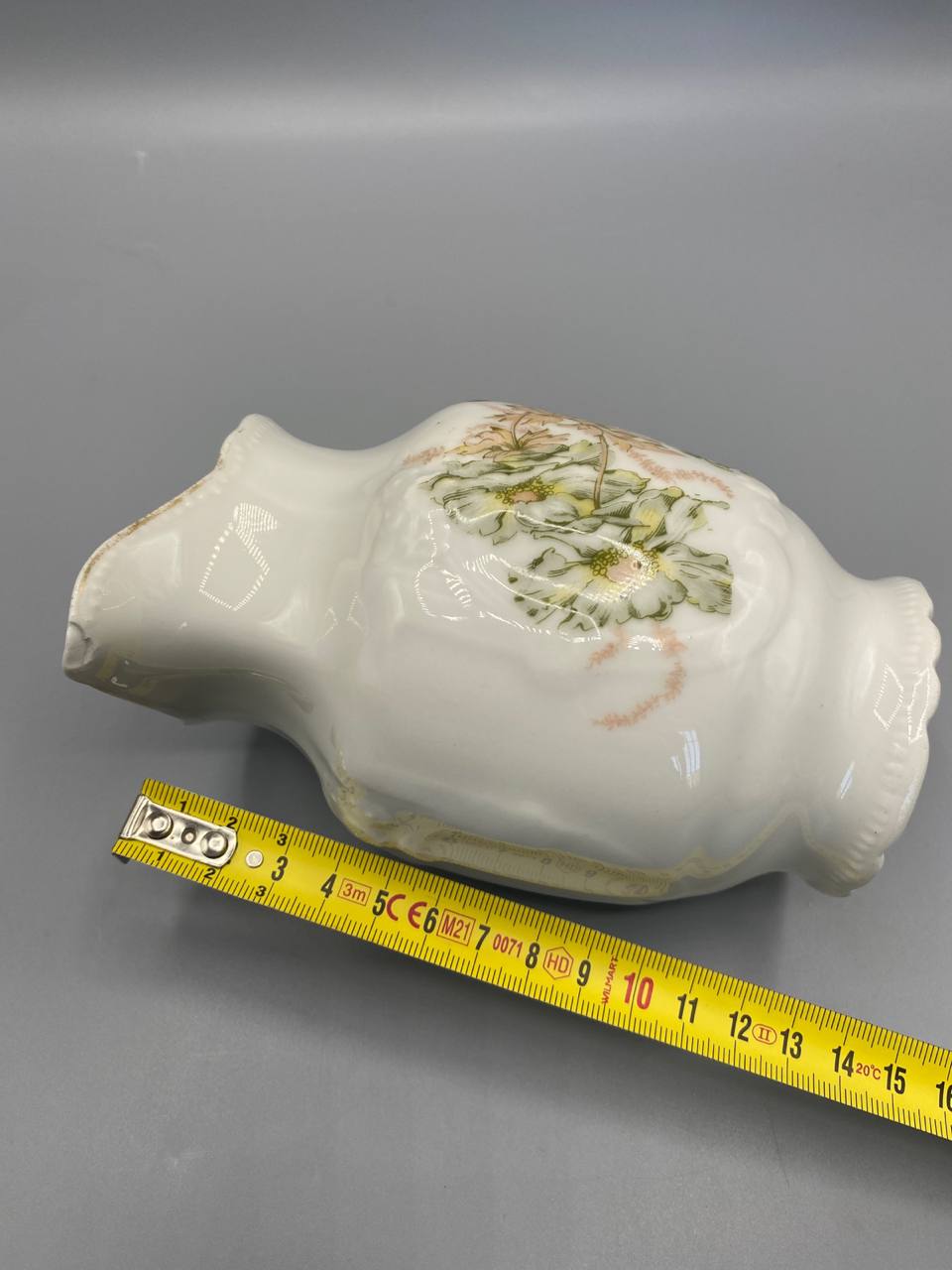 Pot à lait porcelaine couché avec mètre ruban, longueur environ 14 cm, décor floral vert et rose visible sur la panse, fond blanc non émaillé