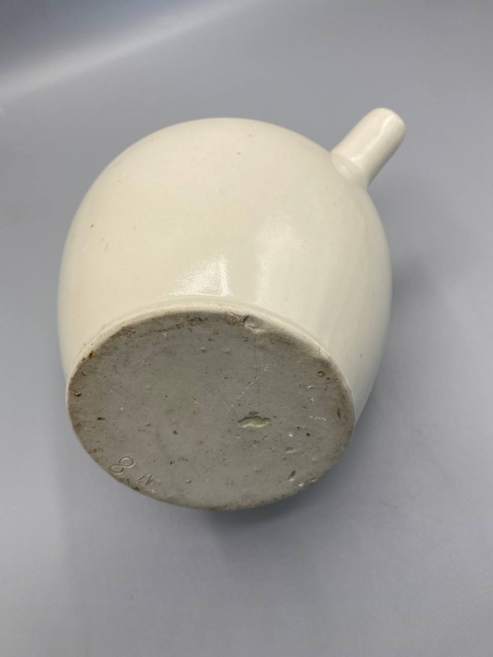 Fond de la gargoulette, pied plat non émaillé en grès gris-beige fin, numéro de moule 84 incisé, bec tubulaire visible en arrière-plan