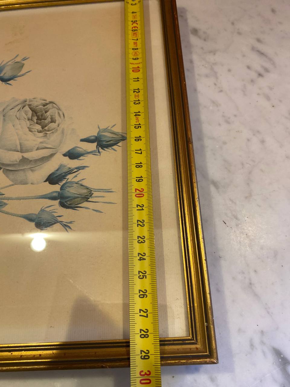 Mesure de la hauteur du cadre avec mètre ruban, environ 50 cm, angle supérieur droit du cadre doré cannelé, rose blanche et boutons bleus visibles
