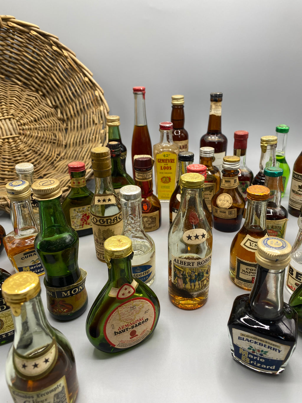 32 mignonnettes anciennes de spiritueux – Vue avec panier en osier plein, De Laroche Cognac et Armagnac au premier plan