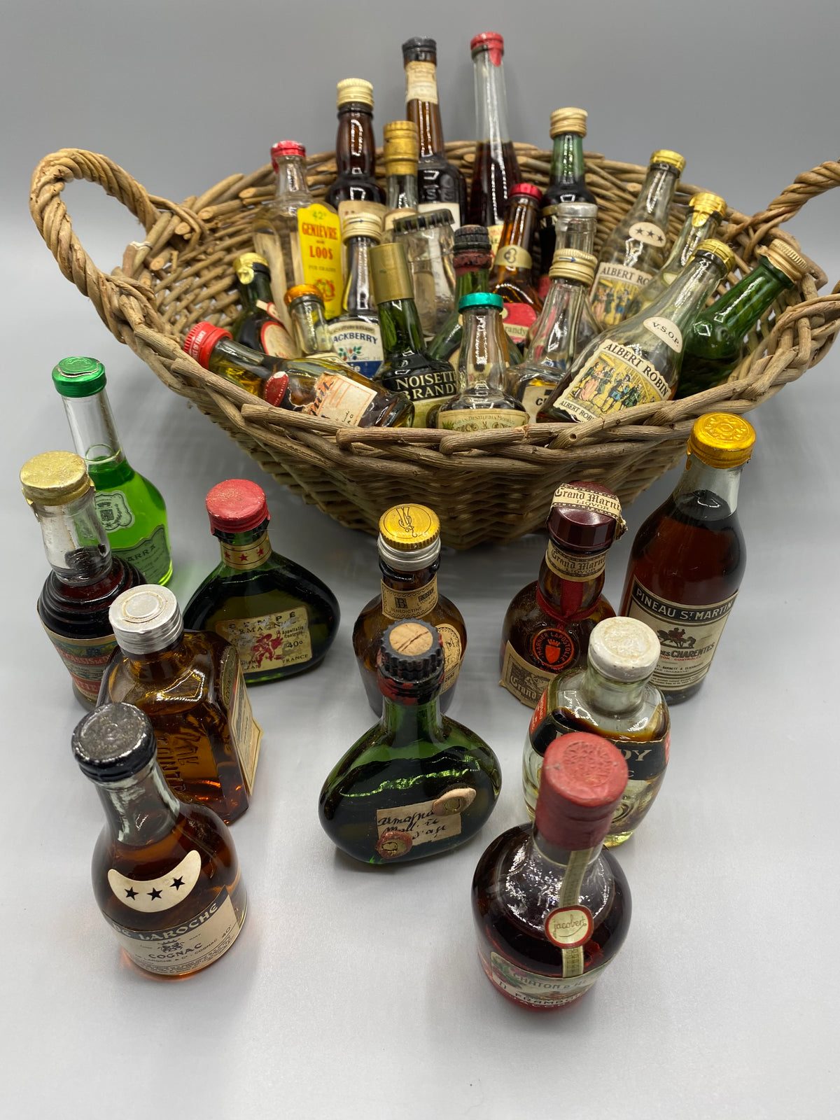 32 mignonnettes anciennes de spiritueux – Vue avec panier et bouteilles autour, Armagnac et Framboise Jacober